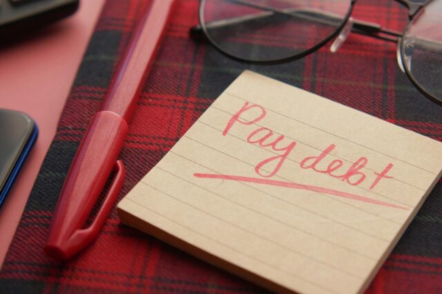 Debt Management calculators_Close-up of a note reading 'Pay debt' next to a red pen on a plaid fabric, emphasizing financial remin